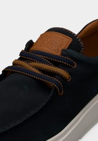 Σκούρο μπλε σουέντ παπούτσι με κορδόνια καφέ και ναυτικού μπλε, δερμάτινο ταμπελάκι ταμπά με ανάγλυφο το όνομα της μάρκας και λευκή καουτσούκ σόλα.