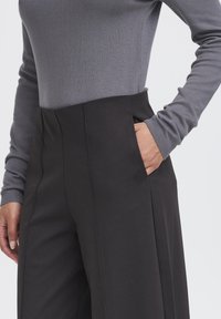 Osoba ubrana w czarne spodnie z wysokim stanem, z ręką w kieszeni, oraz dopasowany, długi szary top na jednolitym tle.