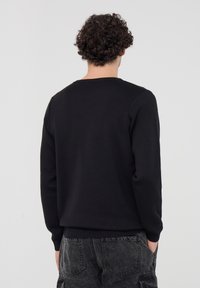 Maglione nero lavorato a maglia, con scollatura rotonda e polsini a coste; indossato con pantaloni scuri a sigaretta, che mostrano una vista posteriore semplice e testurizzata.