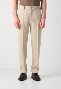 Pantalones de lino beige de corte recto, con cierre de cremallera y botón, textura sutil y bolsillos laterales. Llevados con zapatos negros.