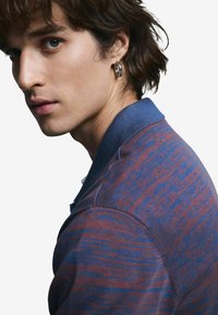 Pánske polokošeľa s textúrovaným vzorom v tmavočervenej a modrej farbe, s kontrastným modrým golierom a detailom jedného náušnice.