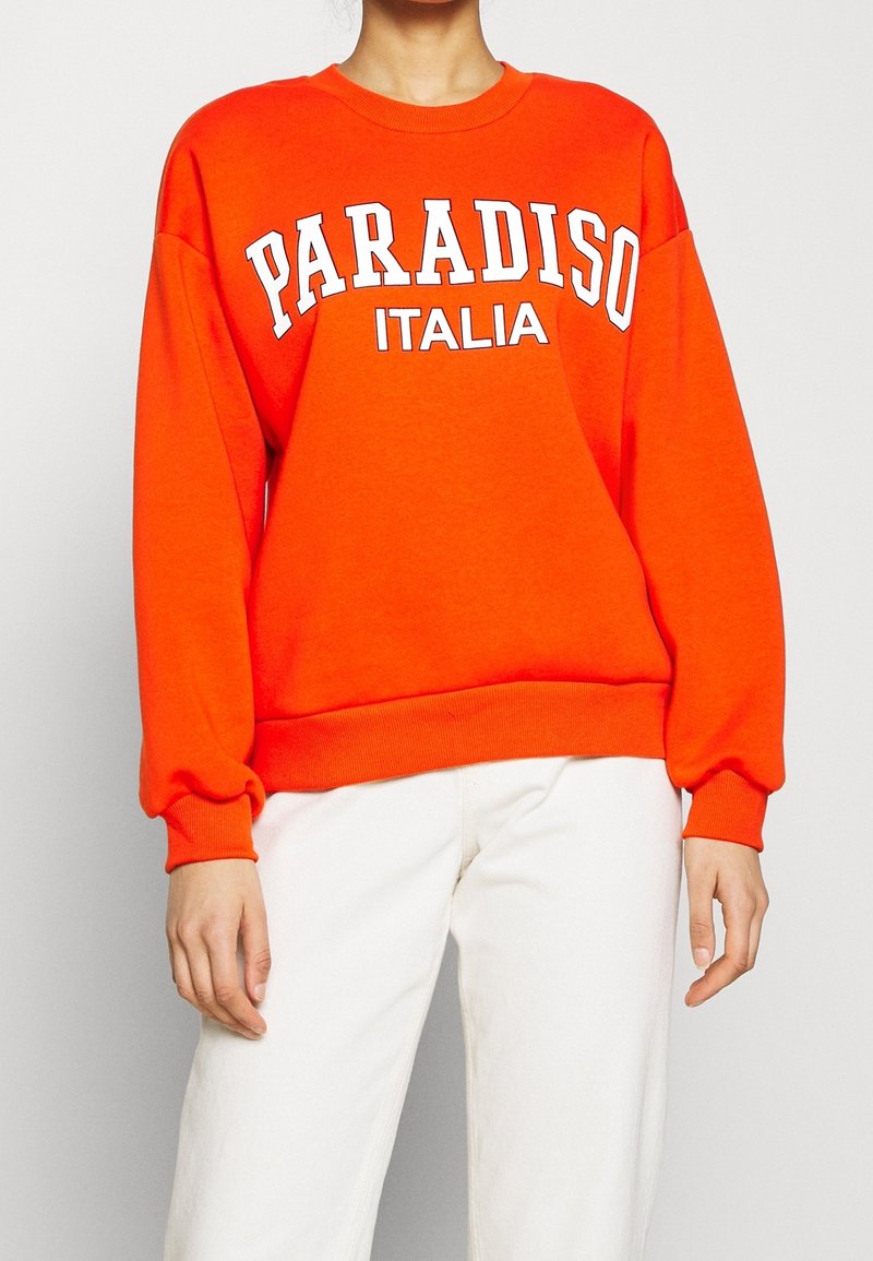 Personne portant un sweat-shirt orange vif avec l'inscription "PARADISO ITALIA" en blanc, associée à un pantalon blanc cassé, debout devant un fond uni.