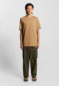 Ljusbrun bomulls-t-shirt, kortärmad, med varumärkeslogotyp på bröstet; kombinerad med olivgröna, avslappnade cargobyxor och mönstrade skor.