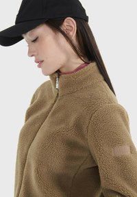 Braune Fleecejacke mit hohem Kragen und Reißverschluss. Verfügt über eine strukturierten Oberfläche und ein Logopatch am linken Ärmel. Getragen mit einer schwarzen Kappe.