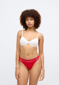 Biały stanik wkładkowy i czerwony koronkowy thong, z cienkimi ramiączkami, gładkim materiałem oraz ozdobnym koronkowym wykończeniem wzdłuż pasa.