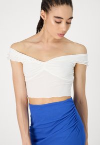 Balta off-the-shoulder apgrieztais tops ar viļņotiem malām un savilktu centru, savienots ar zilu ruched svārku. Gluda auduma tekstūra, pieguļoša dizains.