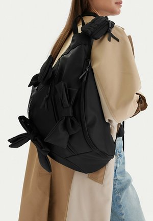 Frau trägt beige Trenchcoat und blaue Jeans, trägt großen schwarzen Rucksack mit mehreren Schleifen- und Reißverschlussdetails.