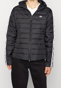 Svart puffjacka med dragkedja och huva. Har horisontell sömnad och vita ränder längs ärmarna, tillverkad av lätt material.