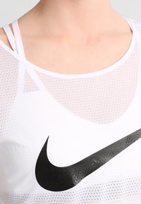 Biały sportowy top na ramiączkach z siateczkową teksturą, z dużym czarnym logo Nike. Widoczne regulowane paski podkreślają jego przewiewny design.