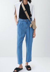 Pantalones de mezclilla de tiro alto en azul claro con pliegues, que cuentan con pasadores para cinturón y un detalle en forma de anillo. Combinados con una blusa negra y sandalias negras tipo slide.