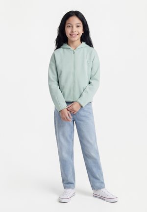 Ung pige med langt mørkt hår smiler, iført en lysegrøn lynlåshættetrøje, lyseblå jeans og hvide sneakers, stående mod en hvid baggrund.