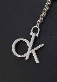 Stříbrný kovový přívěsek ve tvaru "ok" na spojovém řetízku ležící na černém texturovaném povrchu.