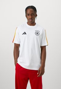 Hvitt kortermet t-skjorte med svart Adidas-logo, Tysklands nasjonalmerke, og rød-gul stripete detaljer på skuldrene.