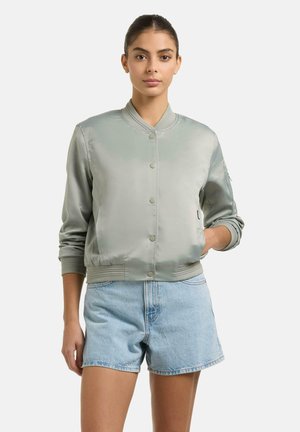 Donna che indossa una giacca bomber in raso grigio chiaro con bottoni a pressione e pantaloncini di jeans azzurri, in piedi con una mano in tasca.