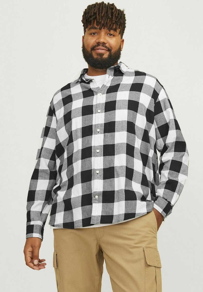 Camisa de franela a cuadros en blanco y negro con mangas largas y botón en la parte delantera, llevada sobre una camiseta blanca, combinada con pantalones caqui.