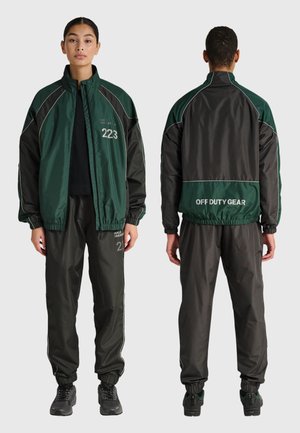 Kvinde iført mørkegrøn og sort træningsdragt med "223" trykt på fronten og "OFF DUTY GEAR" på bagsiden, vist for- og bagfra.