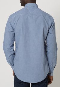 Camisa de botones de cuadros azules y blancos con mangas largas; presenta un cuello extendido y un dobladillo redondeado. La tela parece suave con una textura ligera.