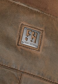 Etichetta in pelle marrone con un logo metallico e lettere in rilievo "FN" e "FREAKY NATION," evidenziate da cuciture a contrasto.