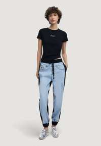 Camiseta negra de manga corta con el texto "Diseñado", pantalones jogger azul claro con paneles laterales negros, cinturilla elástica y zapatillas plateadas.