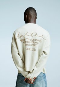 Man met kort haar, gekleed in een crèmekleurige sweater met bruine tekst, met de handen achter zijn rug geklapt, en een losse blauwe spijkerbroek voor een bleke achtergrond.
