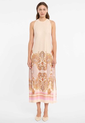 Frau steht und trägt ein ärmelloses, hochgeschlossenes, beiges Maxikleid mit rosa und orangenen floralen und geometrischen Mustern, kombiniert mit beigen spitzen Schuhen.