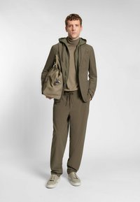Olijfgroene outfit bestaande uit een nauwsluitend langmouwtop, een lichte hoodiejack en bijpassende relaxte broek, gecombineerd met beige sneakers.