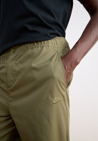 Khaki Nike nadrágok, elasztikus derékkal, oldalsó zsebekkel és arany hímzett logóval, sima, könnyű szövet textúrával.