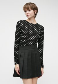 Svart langermet topp med hvite prikker, kombinert med et plissert svart skjørt. Stoffet ser glatt ut; designet er strukturert og klassisk.