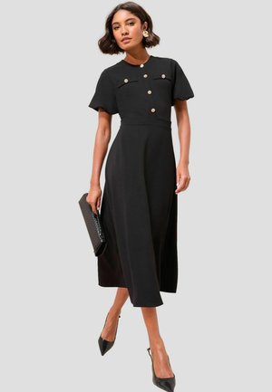 Schwarzes Midi-Kleid mit kurzen Ärmeln, Knopfdetail am Oberteil, zwei vorderen Taschen und glatter Stoffoberfläche; kombiniert mit schwarzen High Heels und einer Clutch.