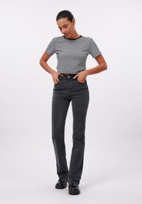 Jauna moteris su tamsiais plaukais, dėvinti dryžuotą trumpomis rankovėmis marškinį ir juodus aukšto liemens džinsus, stovinti su rankomis kišenėse ant baltos fono.