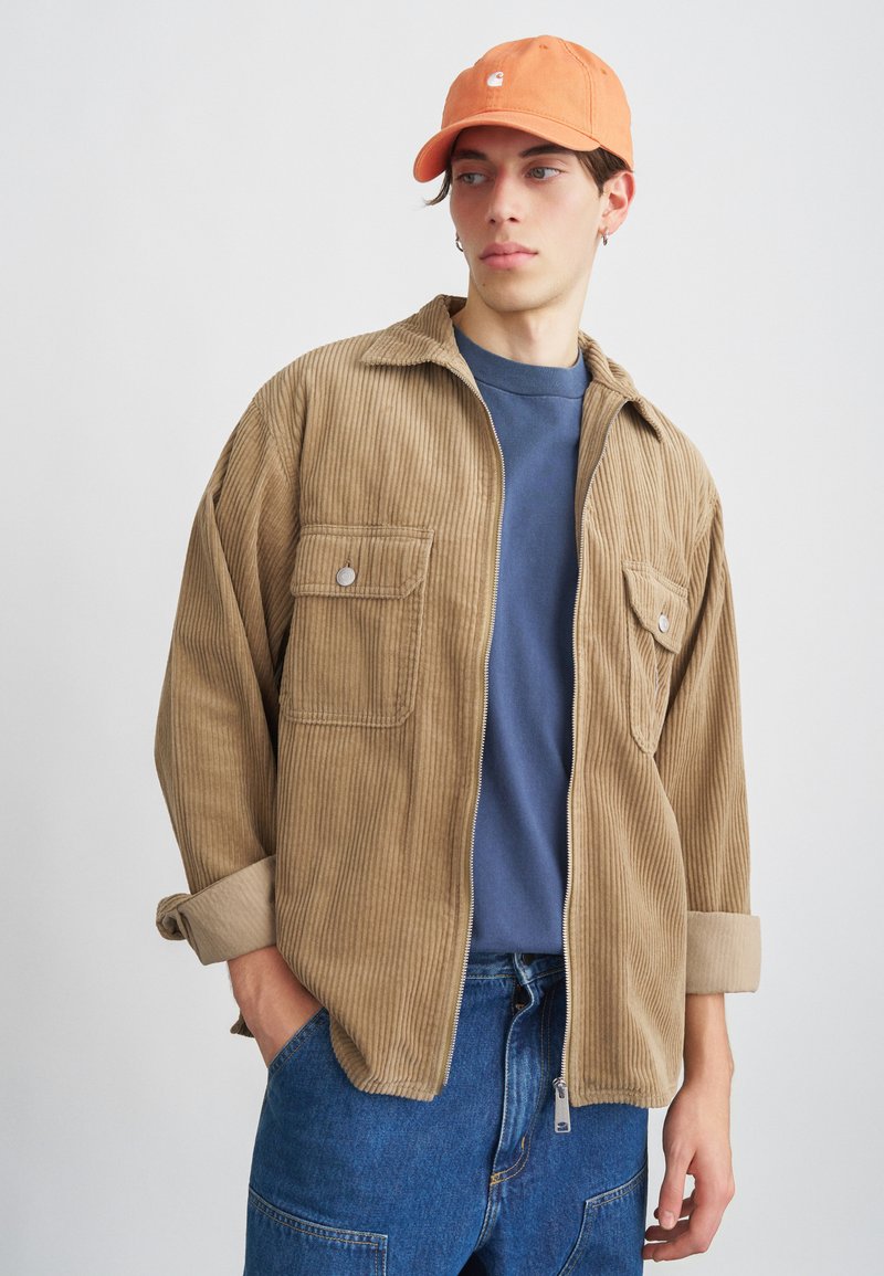Camisa de pana marrón con dos bolsillos frontales, puños beige, llevada sobre una camiseta azul, acompañada de una gorra naranja clara y unos vaqueros.