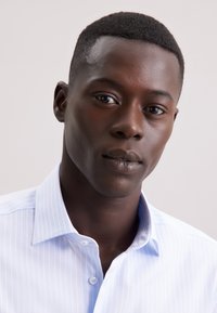 Primo piano di un giovane uomo di colore con capelli corti che indossa una camicia a righe blu chiaro, mentre guarda con calma verso la telecamera.