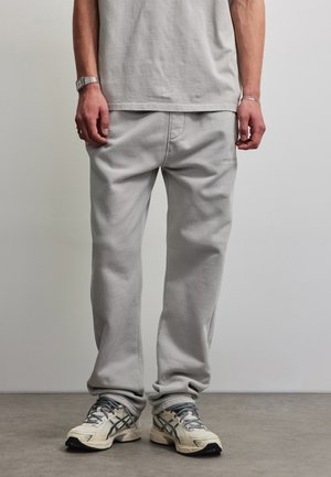Man staande, draagt lichtgrijze joggingbroek, beige en grijze sneakers, een lichtgrijs t-shirt, een polshorloge en een armband.