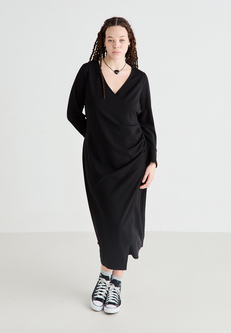 Robe noire à manches longues avec un décolleté en V et un design enveloppant, présentant des détails froncés sur les côtés. Associée à des baskets noir et blanc.