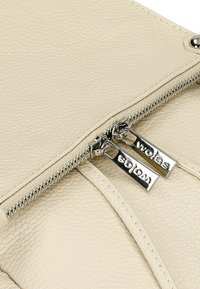 Beige textuur leren tas met een dubbele rits, voorzien van zilverkleurige hardware en gegraveerde labels. Close-up van de ritsdetails en stiksels.