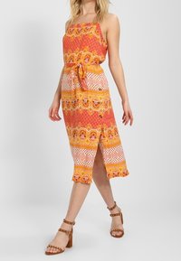 Šaty bez ramínek maxi v živých oranžových a žlutých vzorech s opaskem v pase, rozparkem po kolena a volánovým lemem. Hnědé sandály na podpatku.