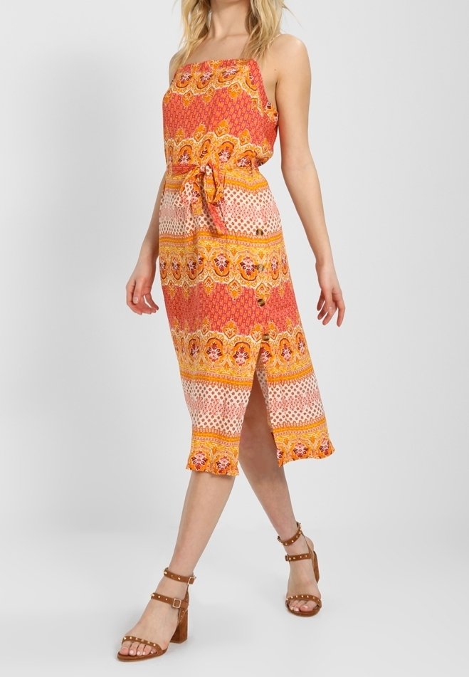 Šaty bez ramínek maxi v živých oranžových a žlutých vzorech s opaskem v pase, rozparkem po kolena a volánovým lemem. Hnědé sandály na podpatku.