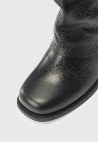 Bota de cuero negro con punta cuadrada redondeada y superficie texturizada sobre un fondo claro y liso.