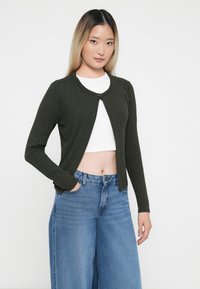 Ribbede grønn cardigan med rund hals og en enkel knappelukking, brukt over en hvit kort topp. Sammen med brede blå jeans.