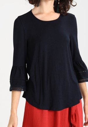 Femme portant un chemisier à manches longues bleu marine avec des poignets en dentelle évasés et un col rond, associé à une jupe rouge.