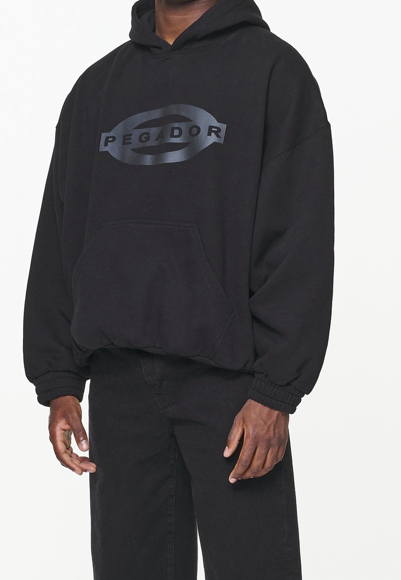 Sweat à capuche noir oversize avec le logo "PEGADOR" sur la poitrine, porté par une personne avec les mains détendues le long du corps, associé à un pantalon noir.
