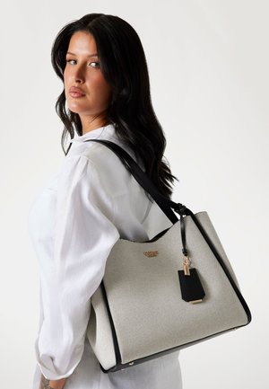 Light gray textured tote bag with a black accent, gold logo, and a detachable tag hangs from a woman's shoulder, featuring dual black handles.