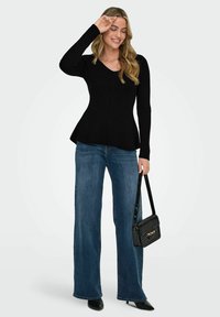 Melna ribota džemperis ar V formas kakla izgriezumu un izplatītu apakšdaļu, kombinēts ar ziliem platām biksēm un melnām šaurā purngala apaviem, tur rokā melnu rokassomu.