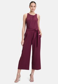 Combinaison sans manches bordeaux avec un design plissé, ceinture nouée à la taille et pantalon large. Le mannequin porte des sandales à talons argentées.