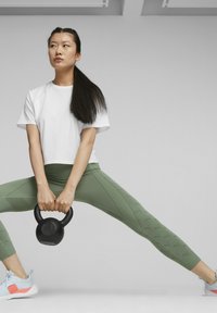 Bijela skraćena majica, zelene teksturirane tajice i svjetloplave atletske cipele. Žena drži crnu kettlebell u poziciji za vježbanje.