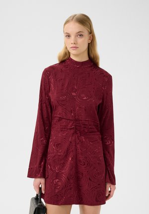 Burgundy mekko, jossa on korkea kaulus, pitkät hihat, teksturoitu kukkakuosi, kerrottu vyötärö ja tiukka siluetti. Mallilla on pieni musta laukku.