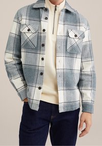 Chemise en flanelle à carreaux bleu et blanc, avec deux poches poitrine, fermeture à boutons et texture douce. Superposée à un pull léger.