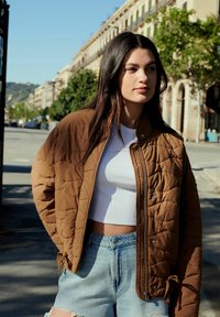 Bruine gewatteerde jas, witte crop top met ronde hals en lichtblauwe denim short. De jas heeft een relaxte pasvorm en wordt in de taille geknoopt.
