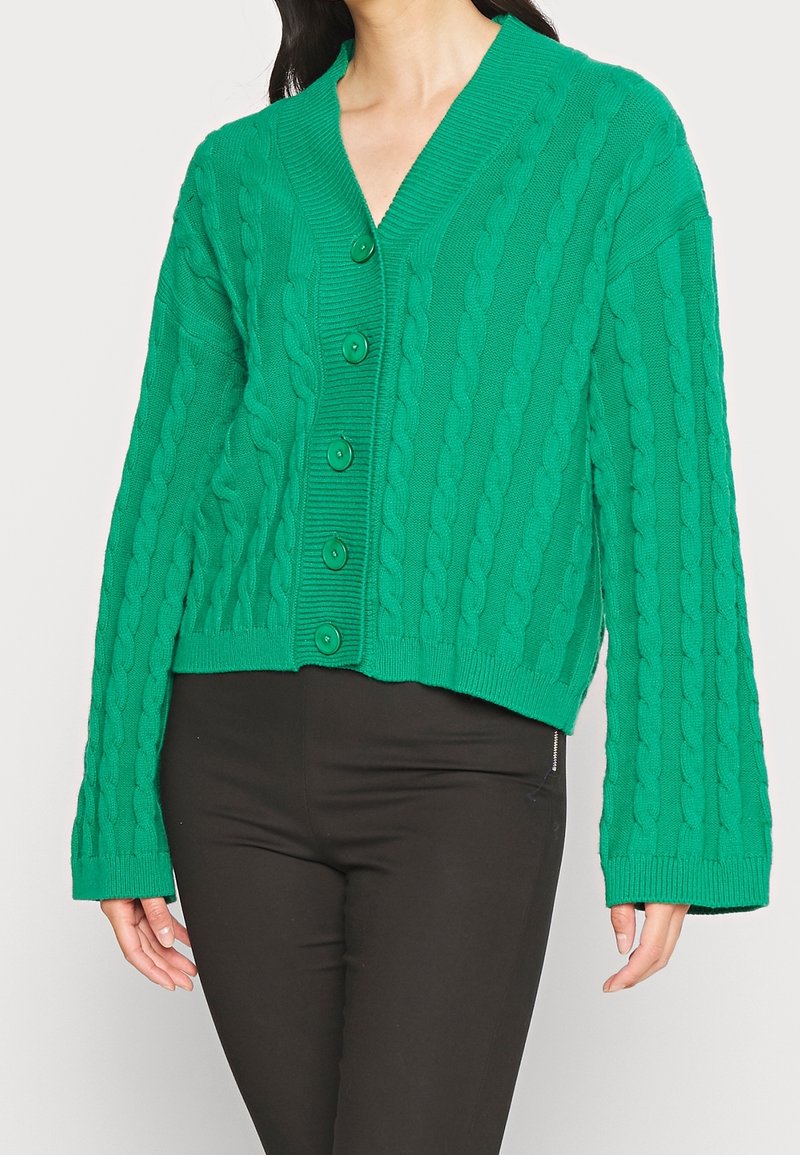 Cardigan vert tricoté à torsades avec un col en V, des boutons sur le devant, et des manches longues et larges. Texture douce avec des accents côtelés au niveau du col et de l'ourlet.