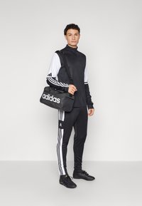 Fekete sportos öltözet fehér díszítéssel, beleértve egy cipzáras felsőt és nadrágot, ami szűkül a bokánál. Fekete sporttáska "adidas" logóval.